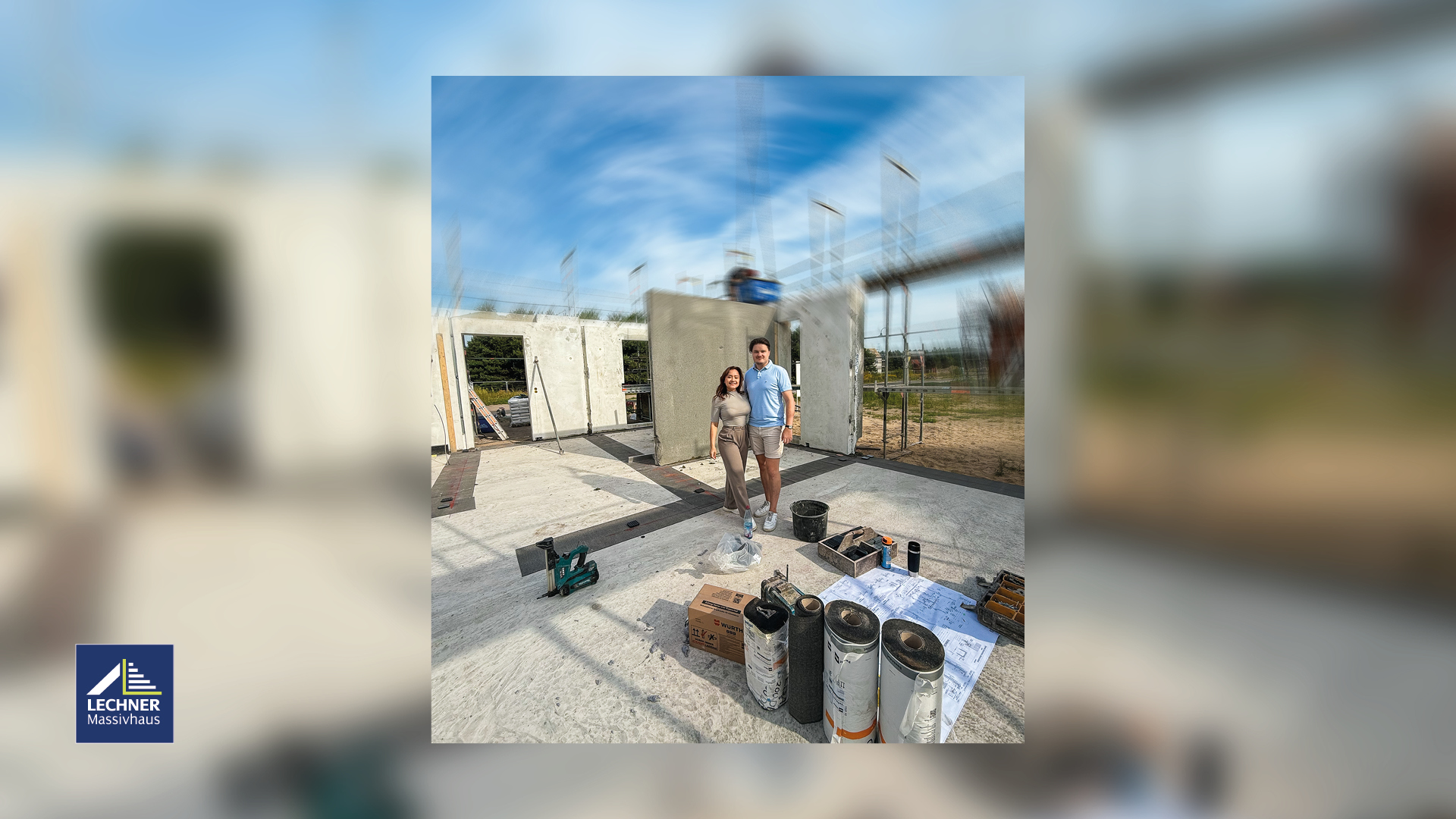 Glückliches Paar vor der Baustelle ihres Massivhauses (Rohbau) von Lechner Massivhaus unter blauem Himmel. Im Vordergrund liegen Baupläne, Dämmrollen und Werkzeug, was den Baufortschritt und die Phase der Bodenplatten-Abdichtung hervorhebt.