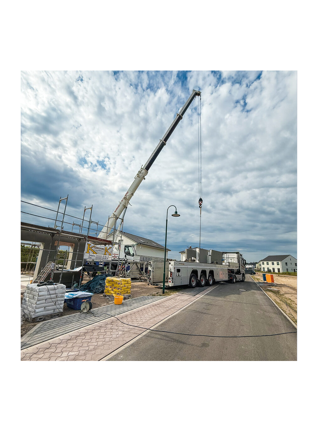 Totale einer Baustelle mit einem großen Mobilkran der Firma K-Kran beim Absetzen von Bauteilen. Auf dem vorbereiteten Bauplatz sind Schalung, Sandsäcke und Baustoffe für den Rohbau von neuen Einfamilienhäusern zu sehen.