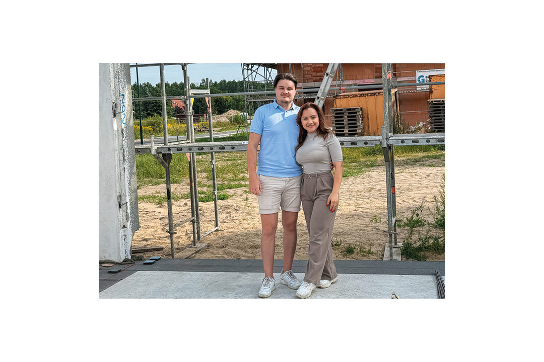 Paar steht lächelnd vor dem beginnenden Rohbau ihres Einfamilienhauses, umgeben von Gerüst und Baufeld. Ein Bild für den Start des Hausbaus und die Freude am Eigenheim