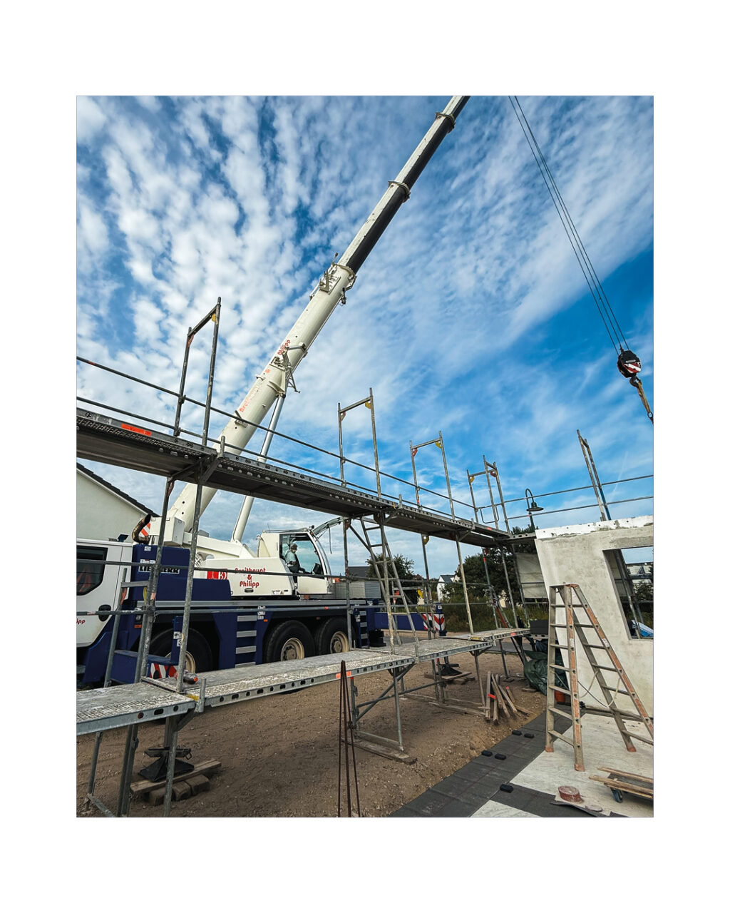 Weitwinkelaufnahme einer Baustelle mit großem Baukran unter blauem Himmel. Das Gerüst des Hauses im Vordergrund und die Maschinentechnik (Kran der P.K.H.Y Firma) symbolisieren den schnellen Fertighaus- oder Massivhausbau.