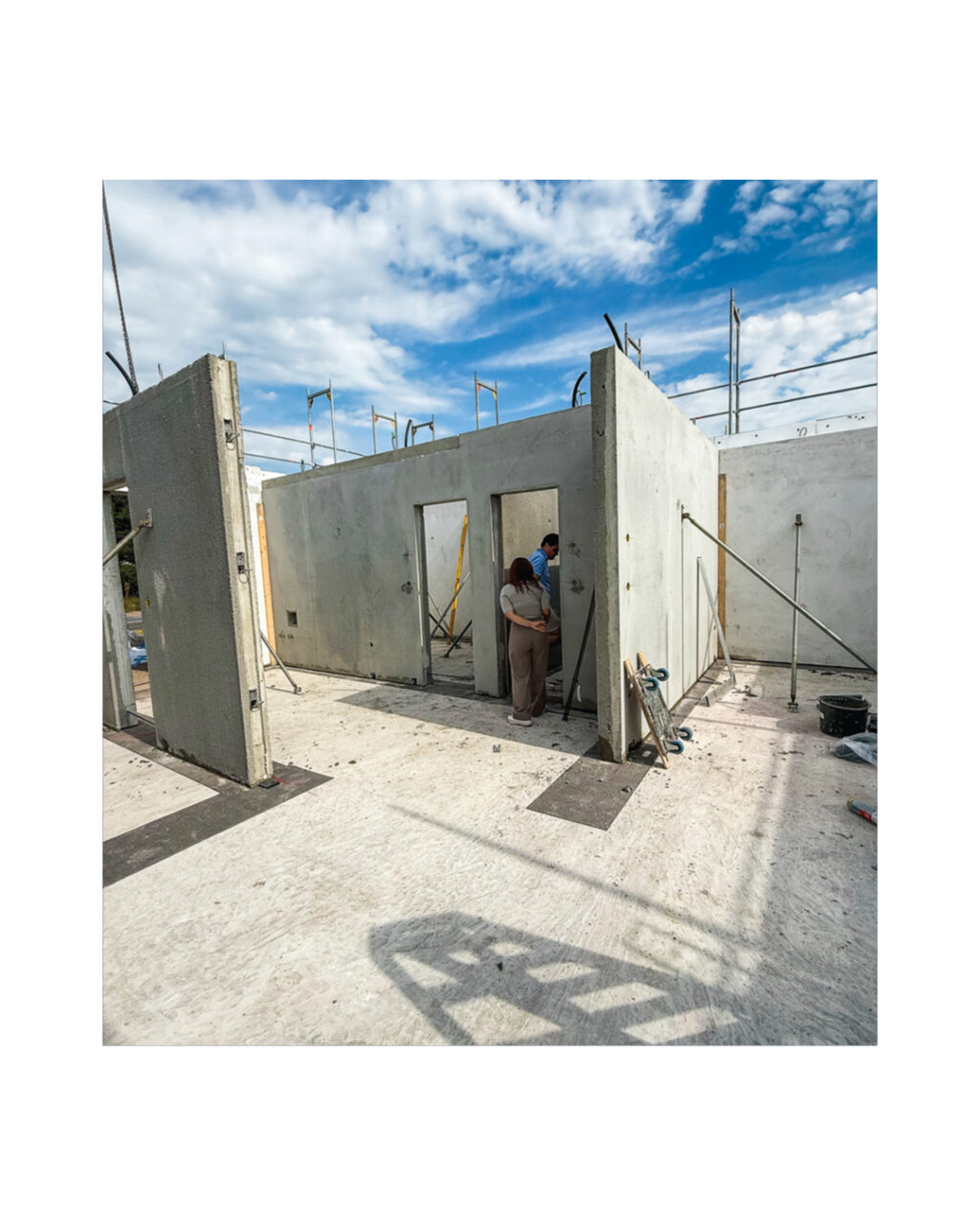 Weitwinkelaufnahme des Rohbaus in der Montagephase. Fertige Betonwände stehen. Bauherren stehen im Türrahmen. Das Foto illustriert den schnellen Baufortschritt und die Errichtung des Massivhaus-Erdgeschosses.