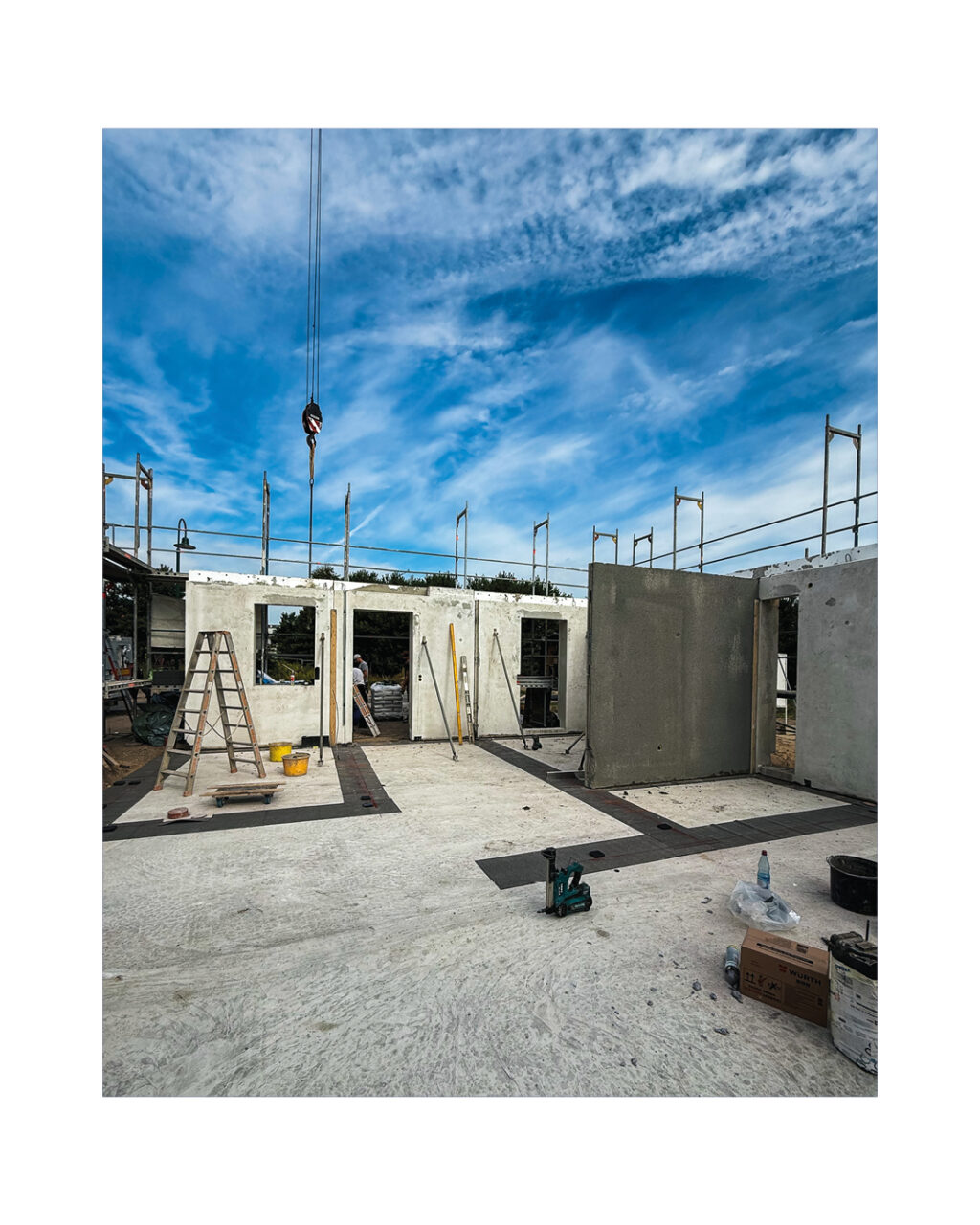 Blick in den halbfertigen Rohbau mit Fensteröffnungen und montierten Betonfertigteilwänden. Ein Kranhaken hängt bereit für die nächsten Wände, was die Effizienz der Massivbauweise darstellt.