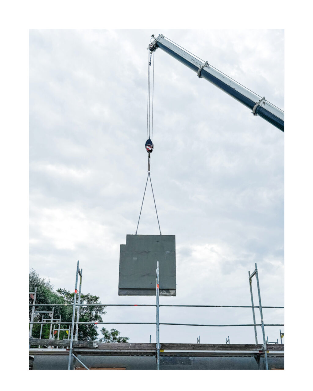 Großaufnahme eines Baukranes, der ein großes graues Betonfertigteil über das Baugerüst hebt. Zeigt die präzise Montage von Massivwänden auf der Baustelle.